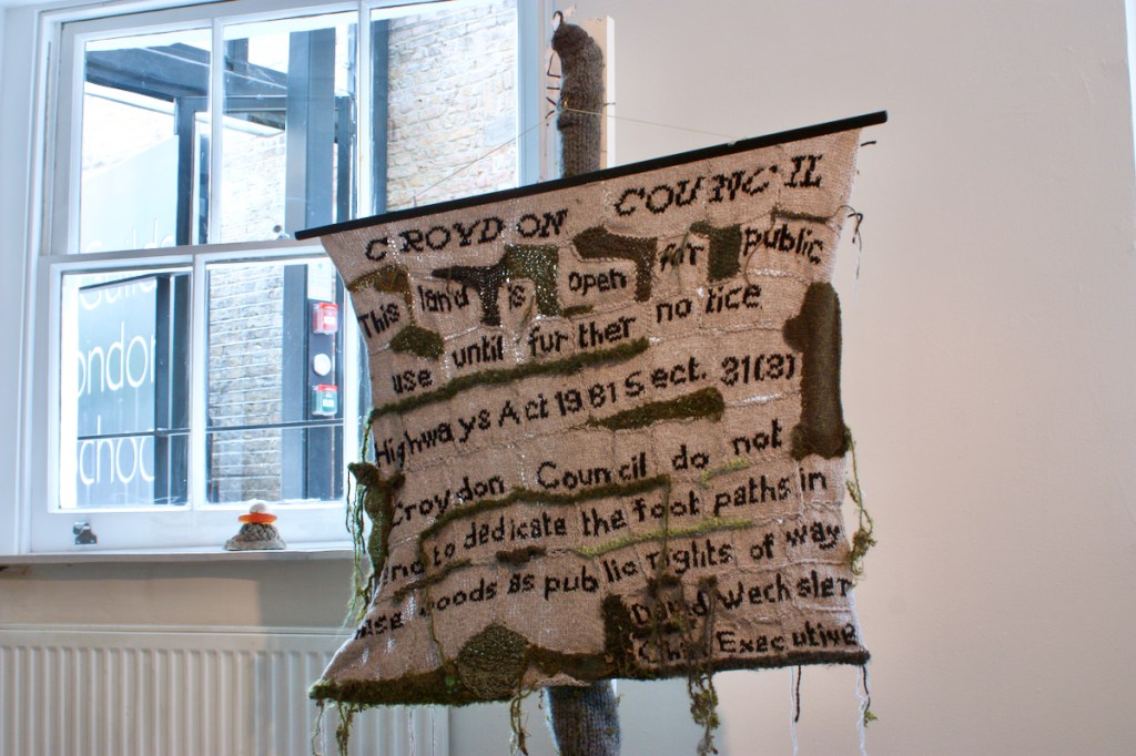 detail of knitted Croydon council sign. Knitted black text on pale grey background saying, 'CROYDON COUNCIL This land is open for public use until further notice. Highways Act 1981 Sect 31(3). Croydon Council do not intended to dedicate the footpaths in these woods as public rights of way. David Wechsler / Chief Executive.' Bits of the words are Interspersed with green mossy patches