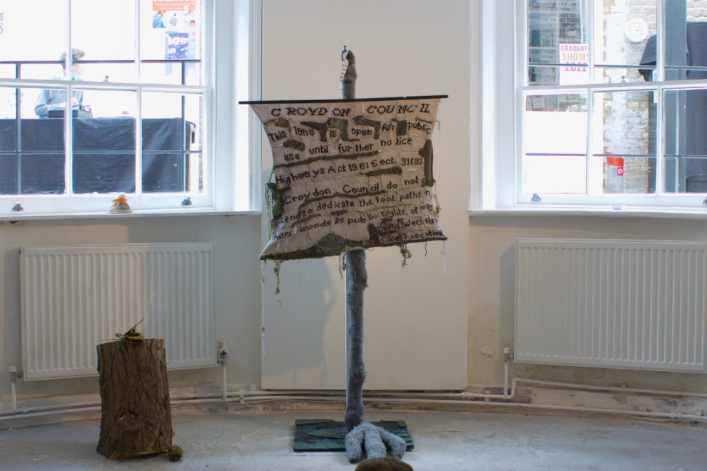 knitted Croydon council sign, the text hangs from poster hangers attached to a chunky grey knitted pole which terminates in a clawed foot.