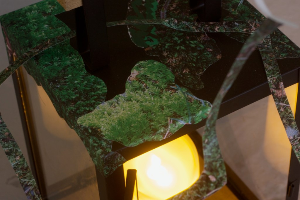 Detail of rectangular black lantern decoupaged with moss images. A LED candle glows inside