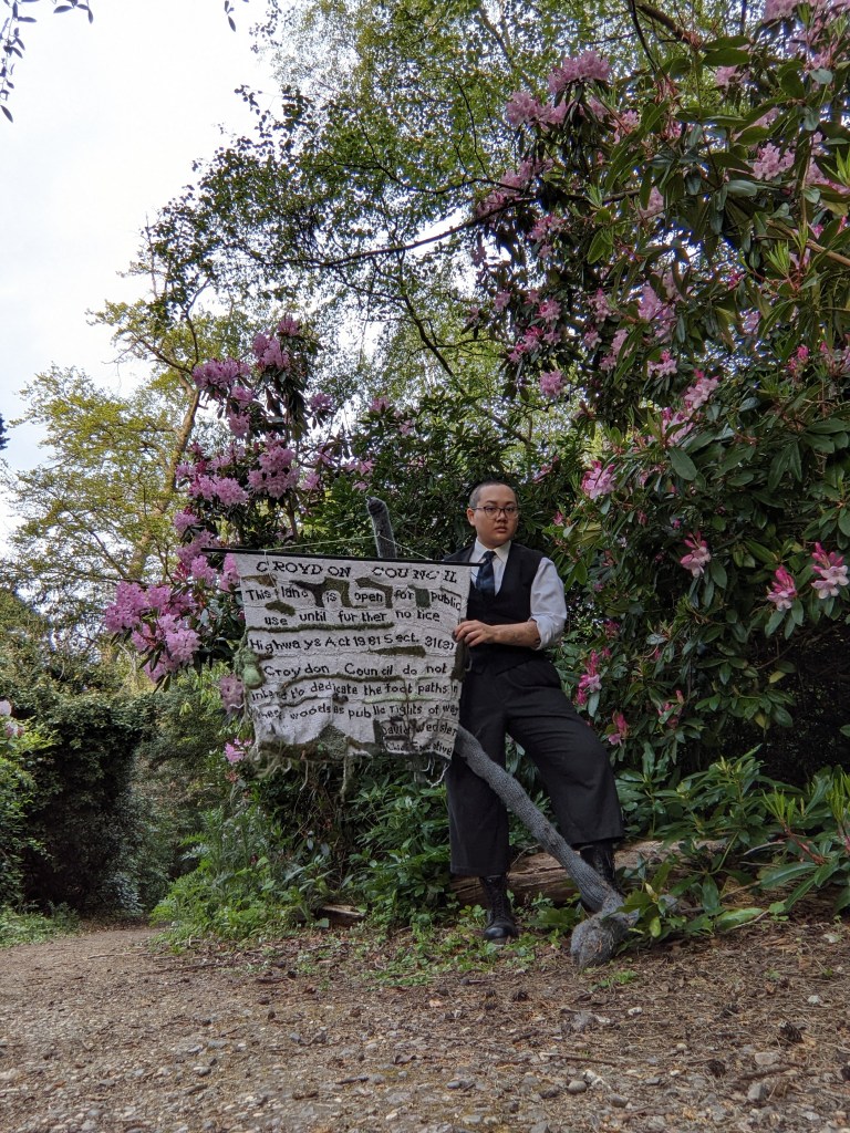 Small bald Asian person in dark waistcoat, trousers, and boots, standing with the knitted Croydon Council sign in front of some rhododendrons. Image taken at Heathfield House, Croydon.